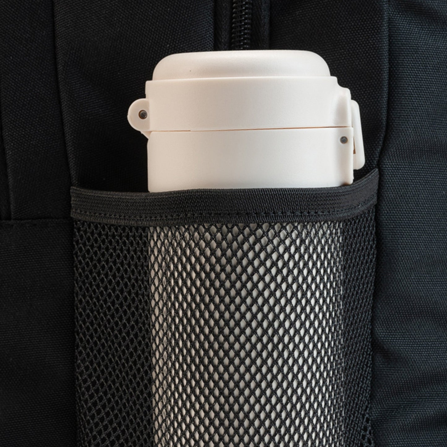 Black backpack side mesh water bottle pocket holding a white travel cup with flip lid, close-up detail view