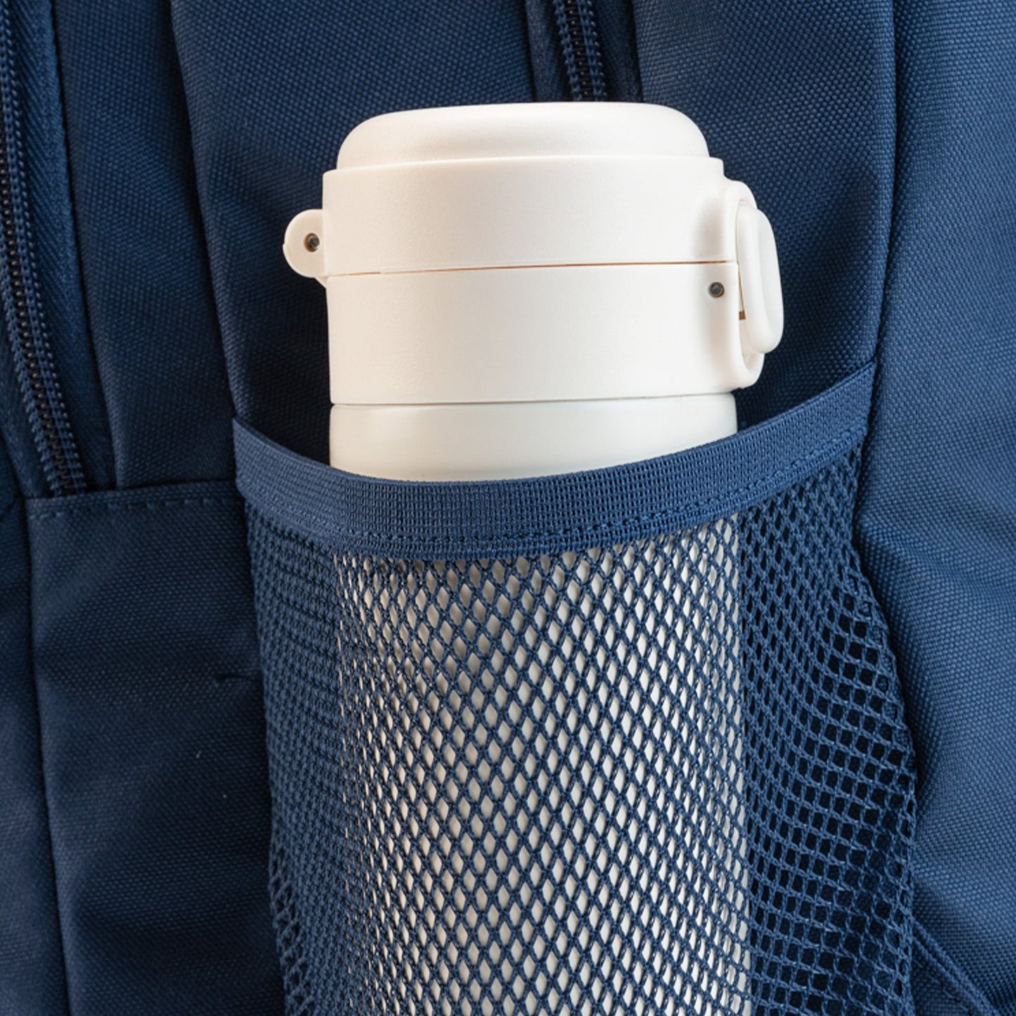 Navy blue backpack side mesh water bottle pocket holding a white insulated flask with clip-lock lid