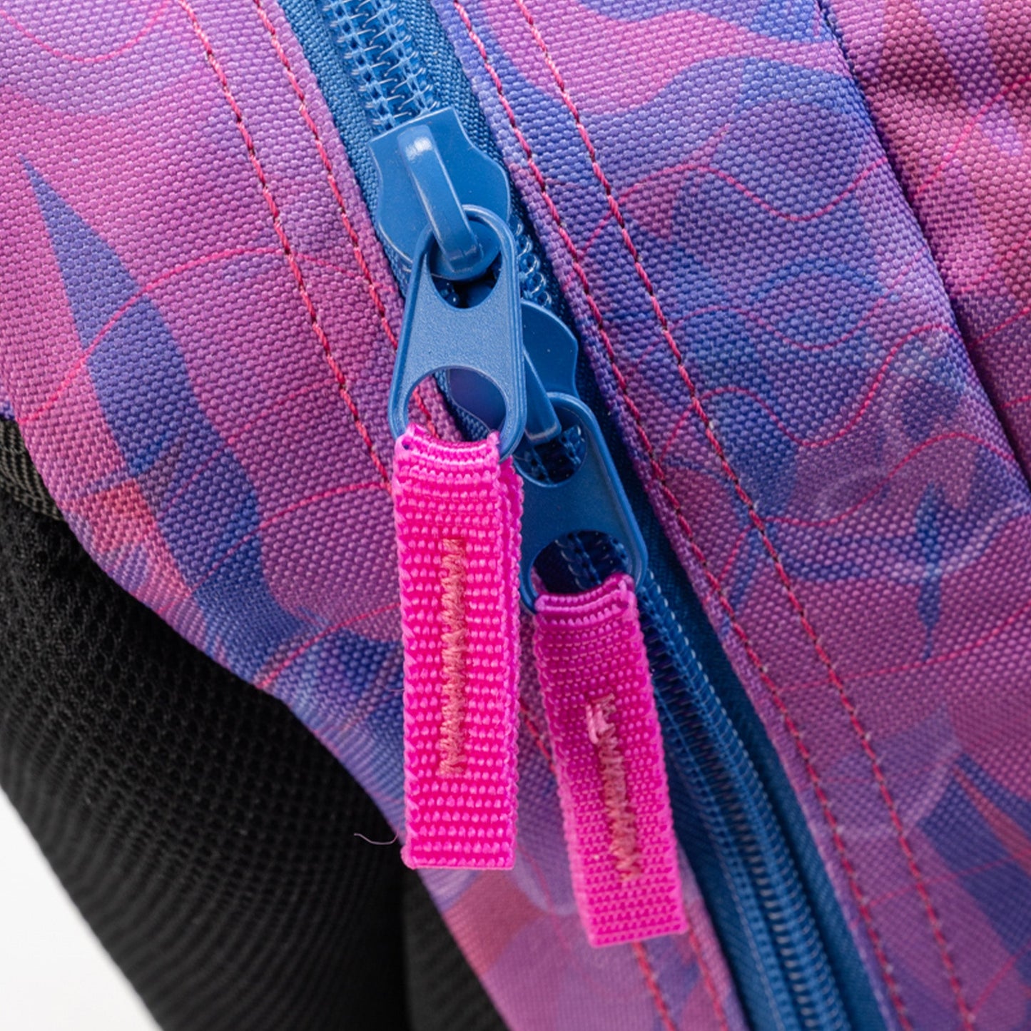 Backpack close-up showing dual blue zip pullers with pink fabric zip tabs on purple and pink geometric print polyester fabric
