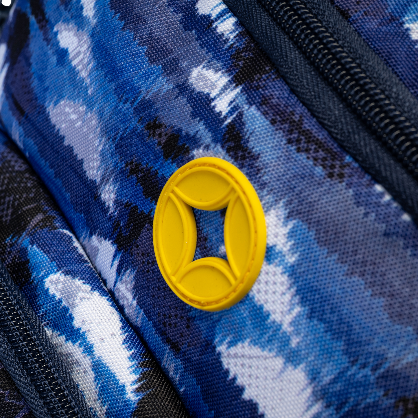 Backpack blue tie-dye camo fabric close-up with yellow oval rubber logo badge and black zipper detail