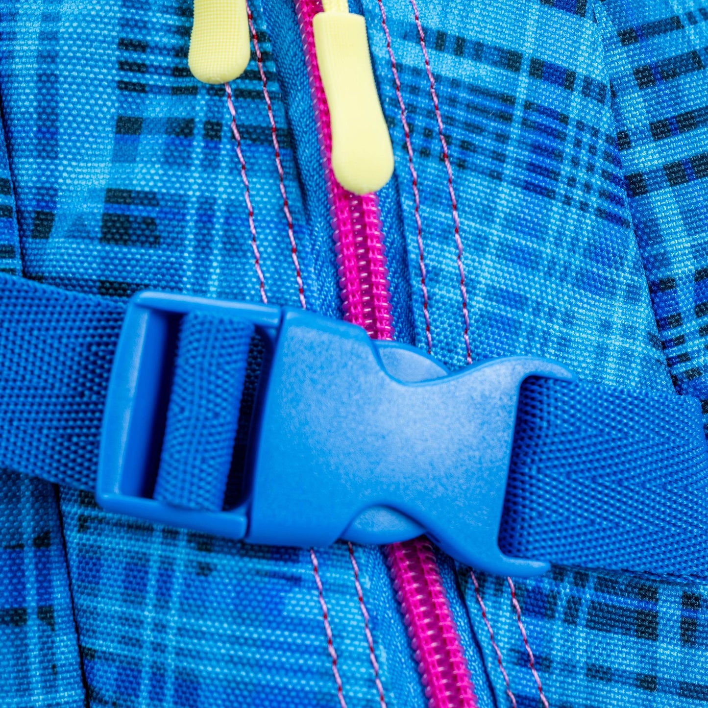 Backpack close-up showing blue plaid nylon fabric, plastic side-release buckle, pink zipper and yellow zip pulls