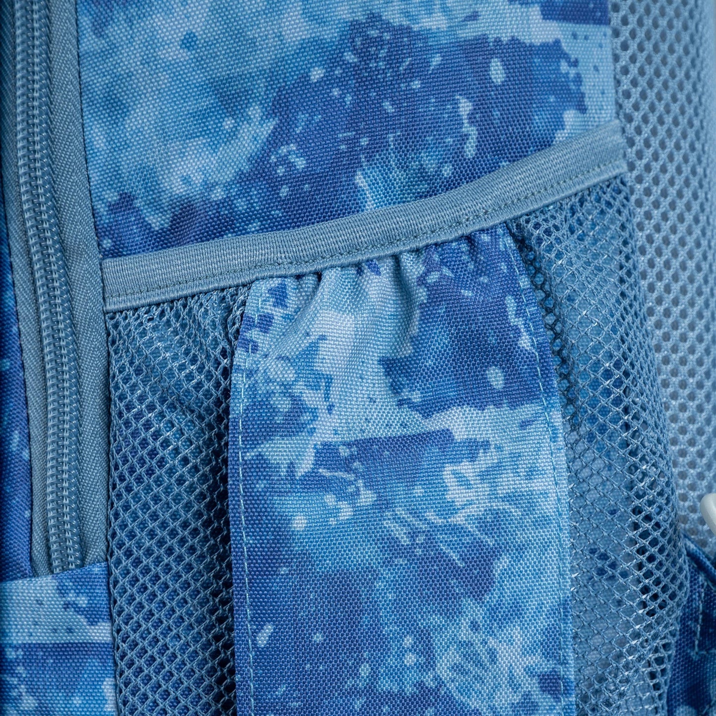 Backpack close-up showing blue tie-dye print fabric, grey mesh side panel, and zip detail with grey binding trim