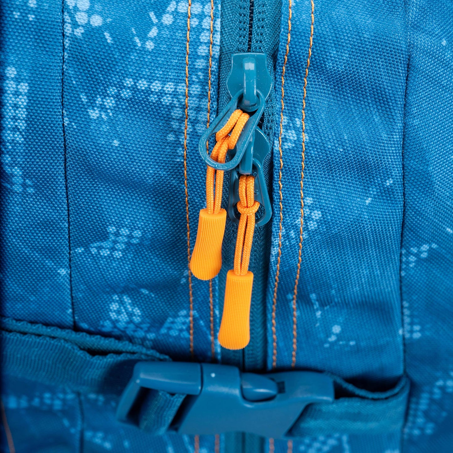 Backpack close-up showing dual zipper pulls with orange cord pulls on blue patterned nylon fabric with orange stitching and teal buckle