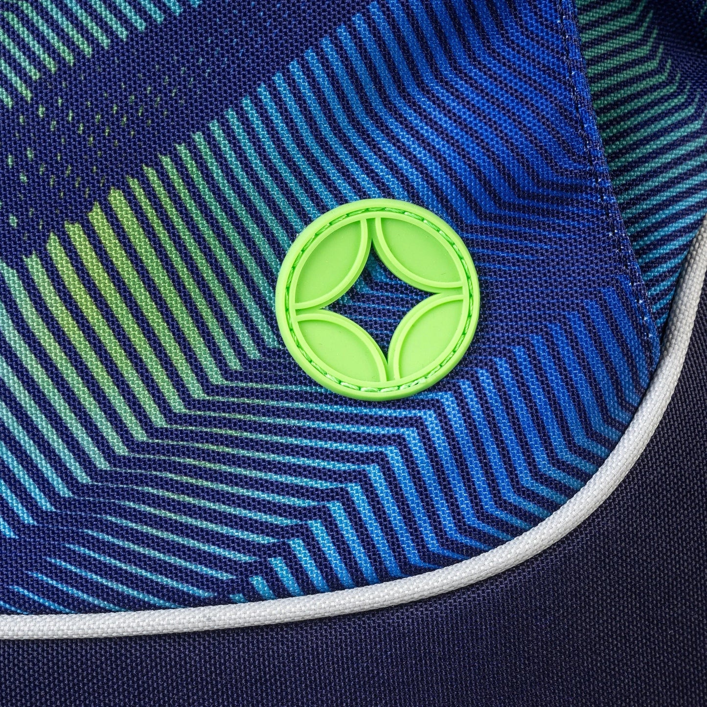 Close-up of bag panel in blue and green geometric zigzag print with neon green circular rubber logo badge and white piping trim