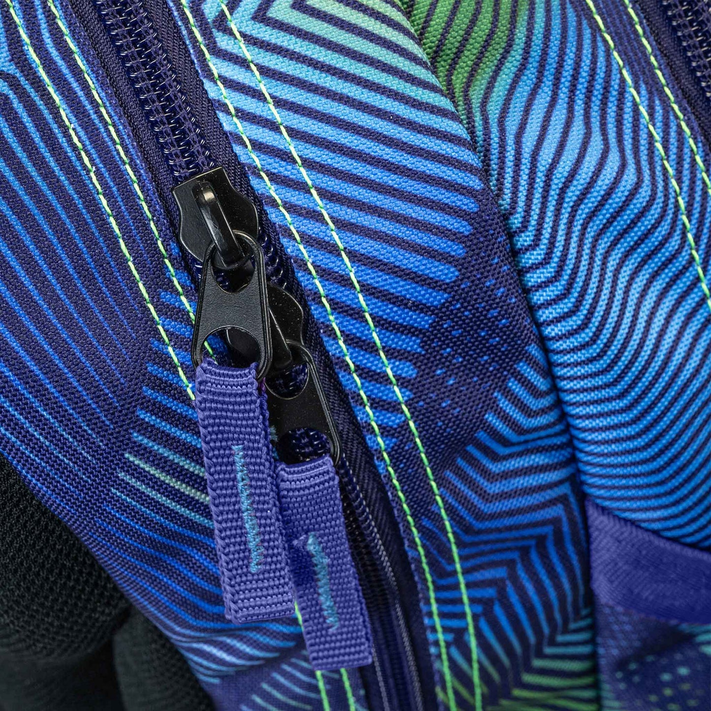 Backpack close-up showing dual black zip pullers with purple fabric tabs on blue green geometric stripe patterned fabric with neon stitching