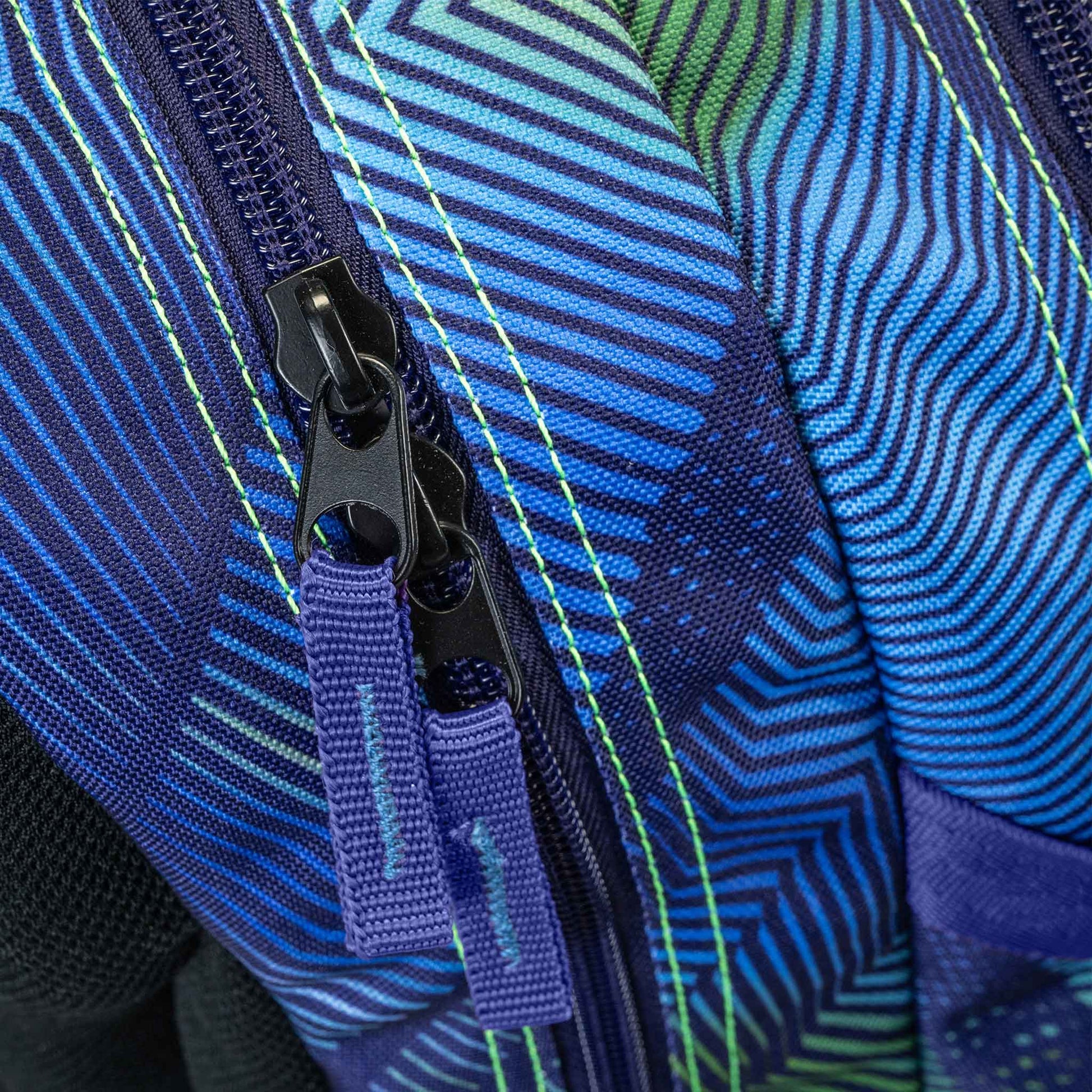 Backpack close-up showing dual black zip pullers with purple fabric tabs on blue green geometric stripe patterned fabric with neon stitching