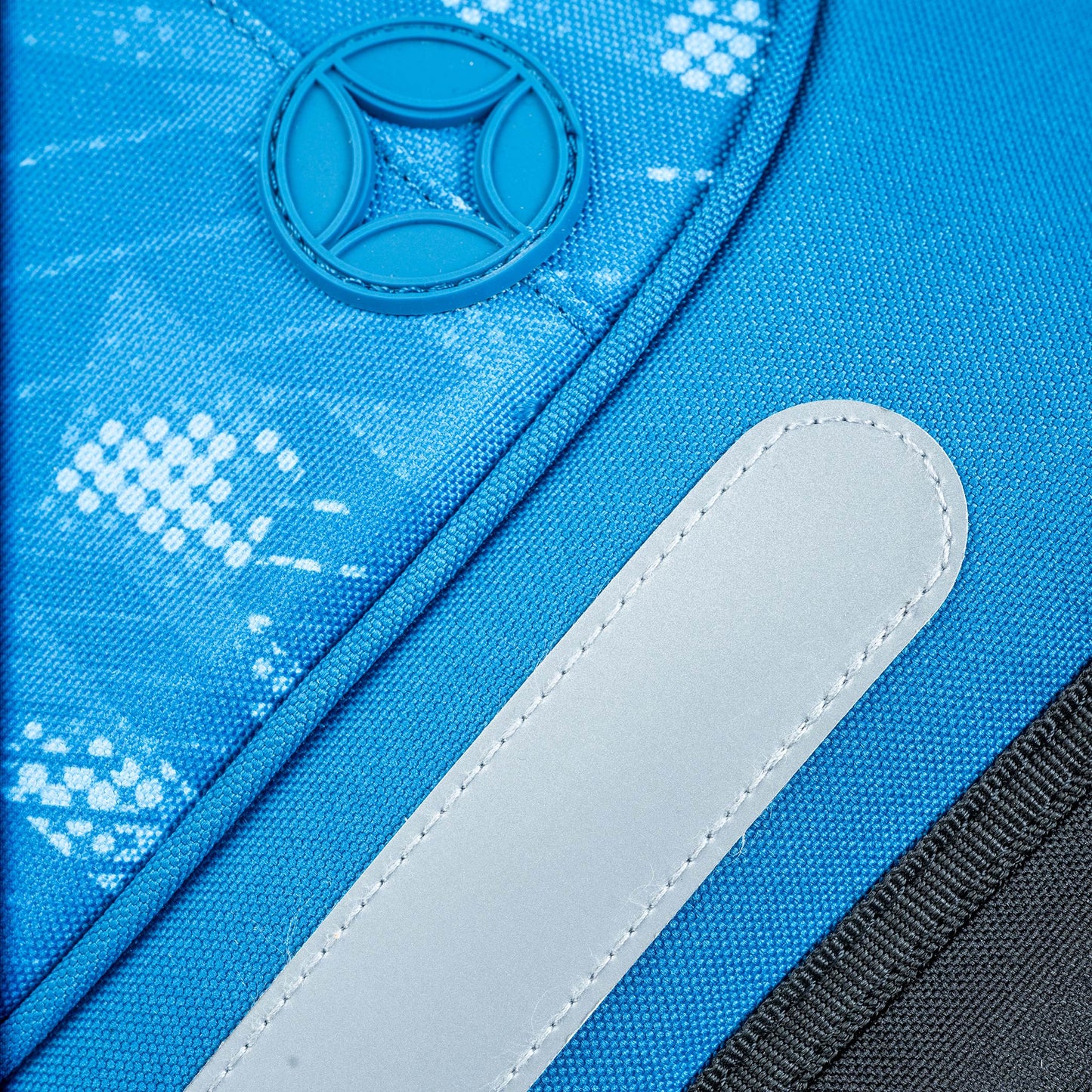 Blue polyester backpack close-up showing white speckle print, rubber logo badge, reflective strip and black zip trim