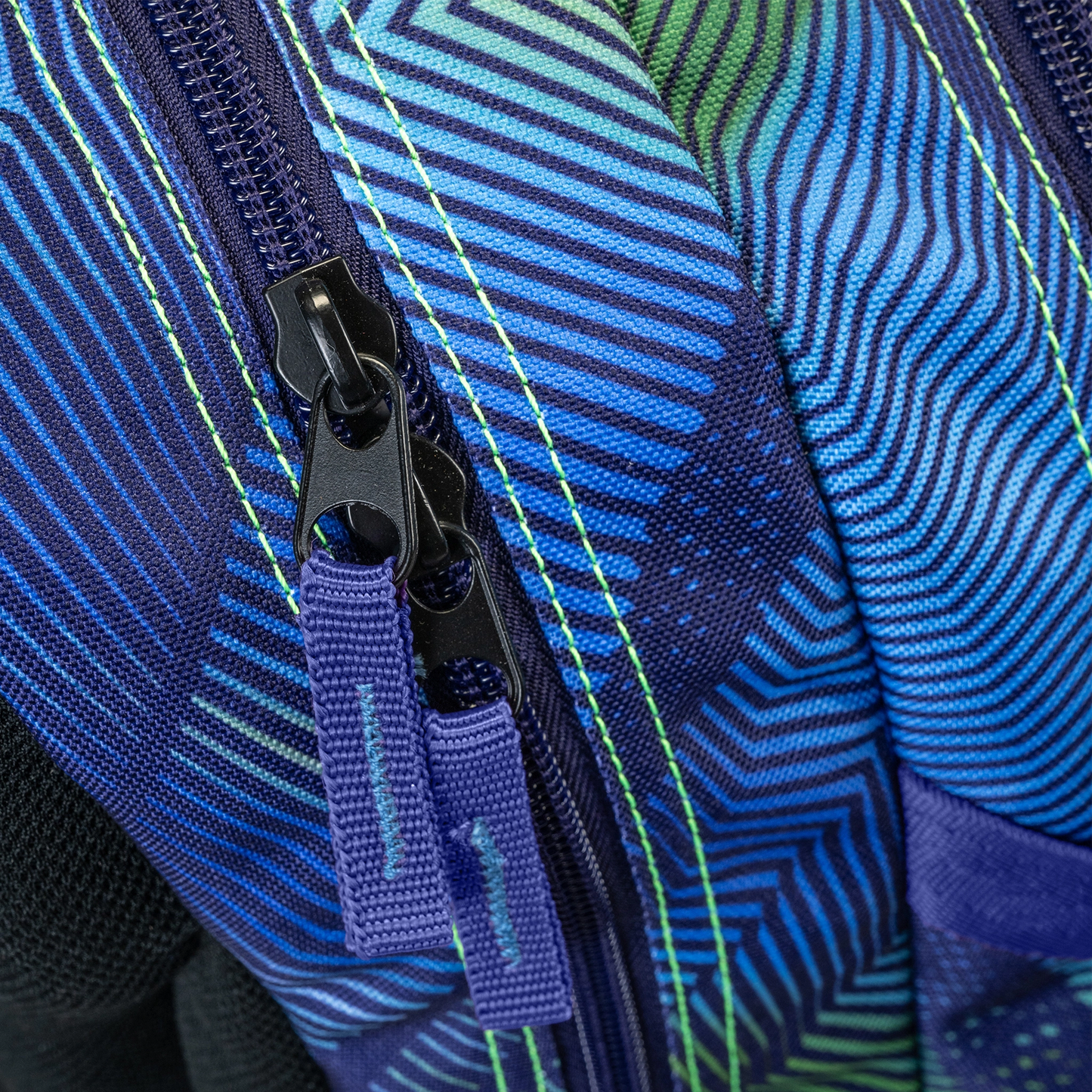 Backpack close-up showing dual black zipper pulls on blue green geometric stripe patterned fabric with neon yellow stitching