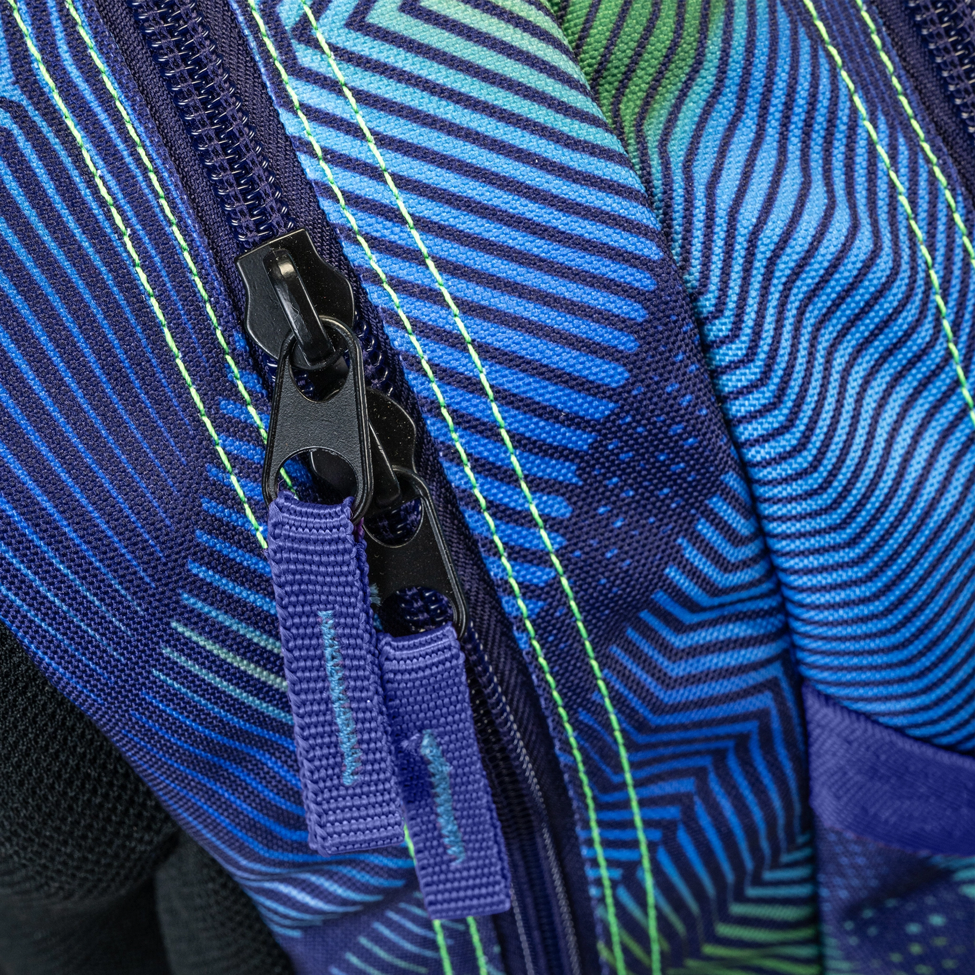 Backpack close-up showing dual black zipper pulls on blue green geometric stripe patterned fabric with neon yellow stitching