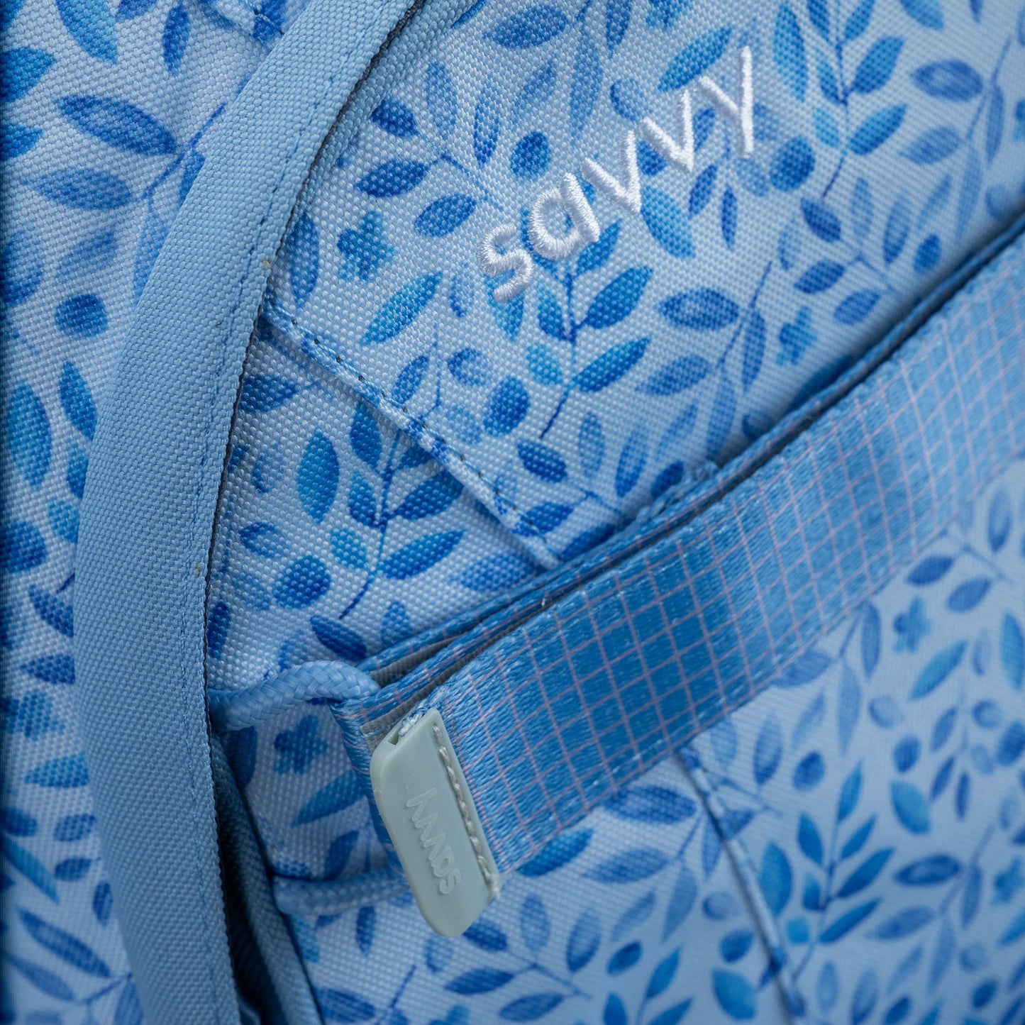 Blue fabric backpack with all-over botanical leaf print, close-up showing zip pocket and embroidered logo detail