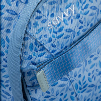 Blue fabric backpack with all-over botanical leaf print, close-up showing zip pocket and embroidered logo detail